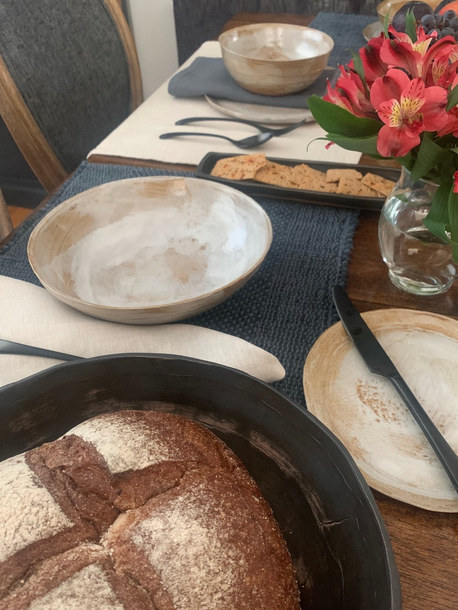 Loaf of bread in a handmade ceramic bowl on a table with flowers, and other items.