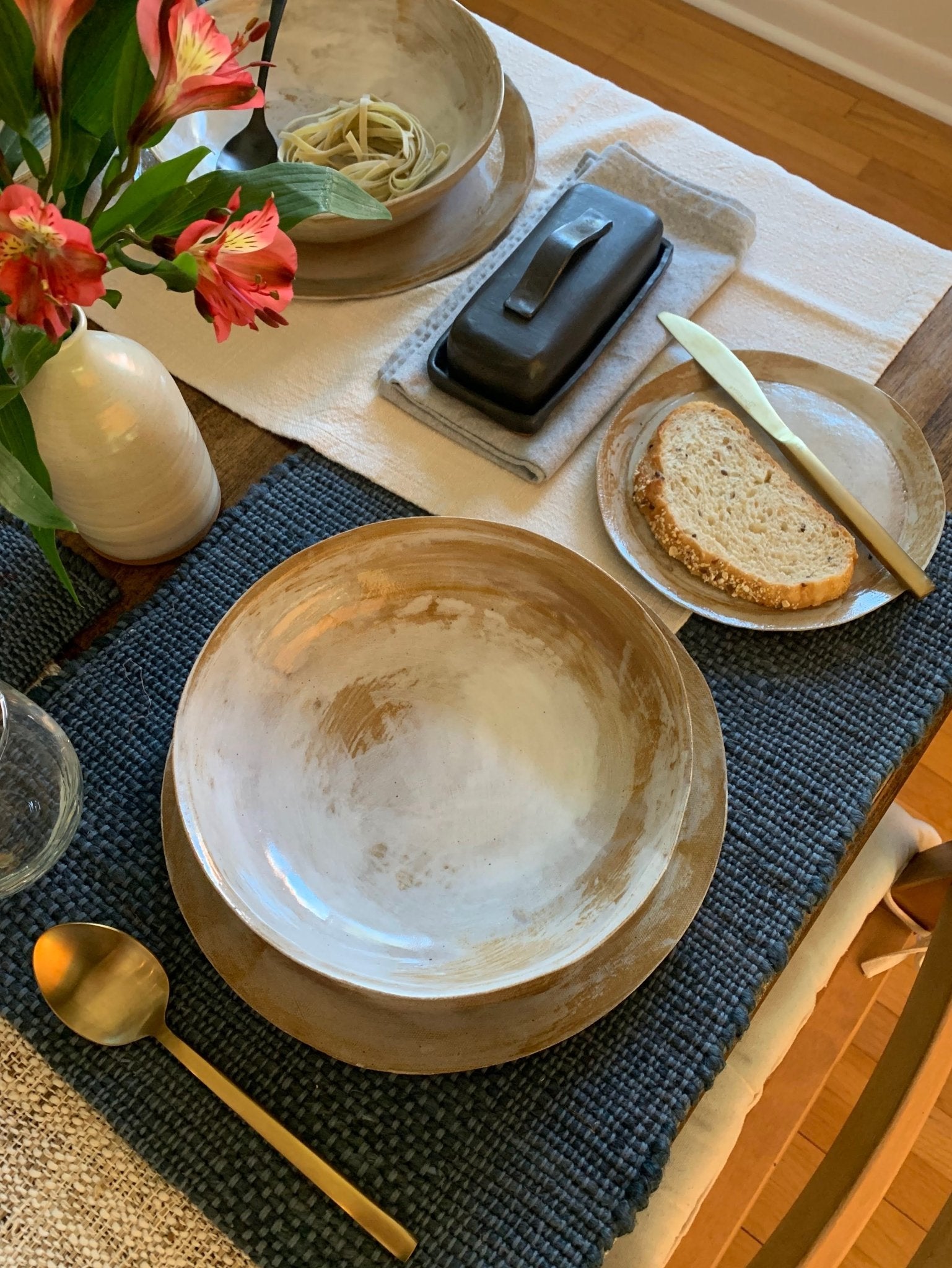 Dining table setting with handmade stoneware, bread, and flowers on a blue placemat.