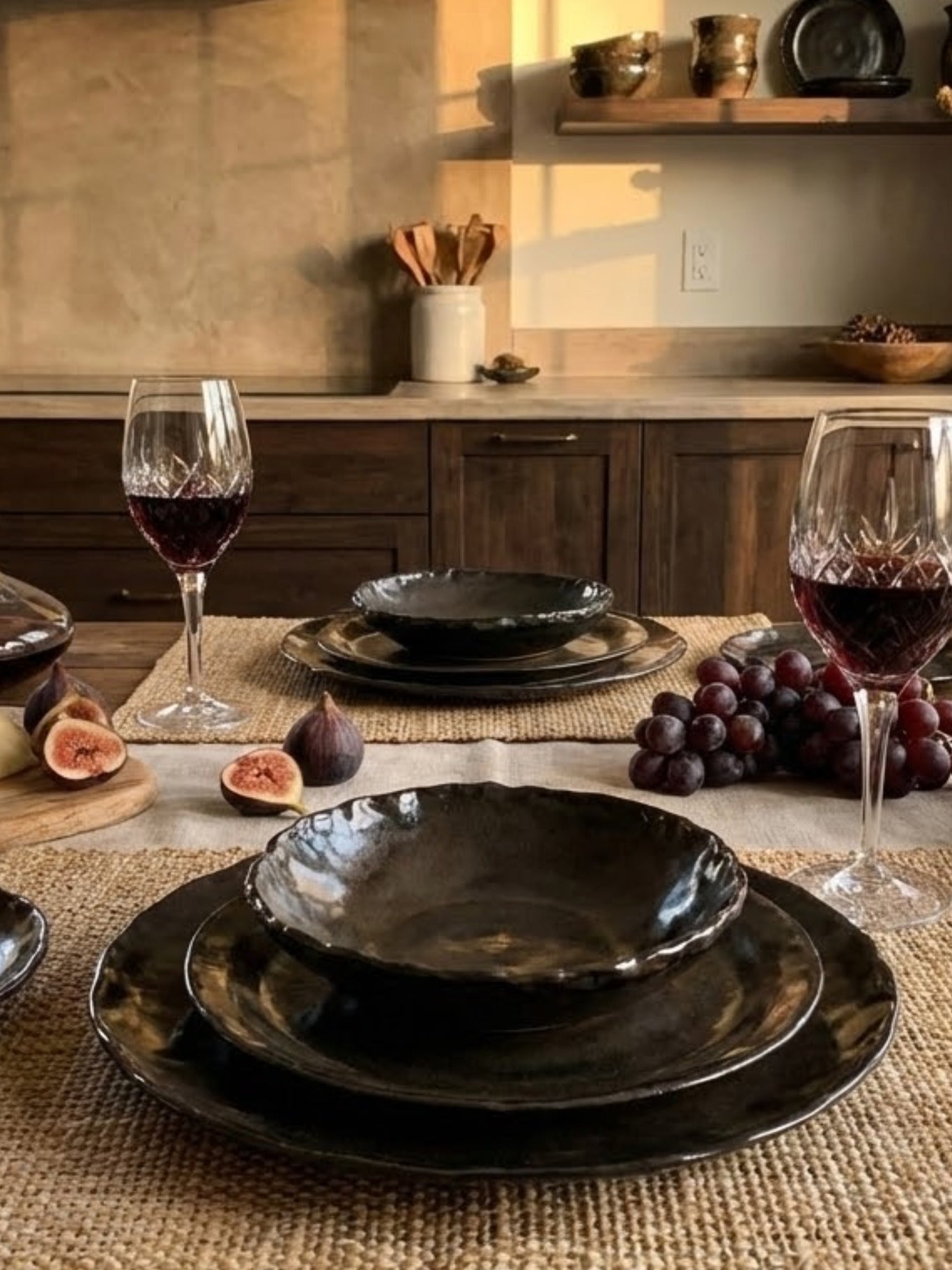 Dining table setting with handcrafted ceramic plates, red wine, figs, and grapes in a warm-toned kitchen.