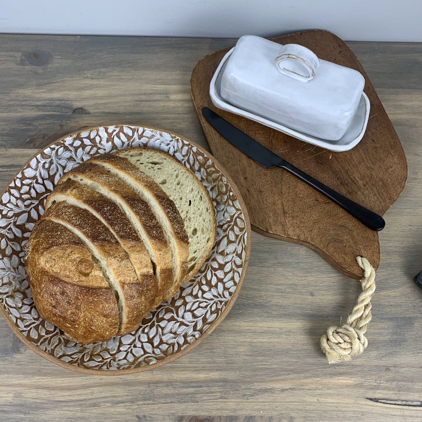 Loaf of bread on a decorative stoneware plate with a wooden cutting board, butter dish, and knife on a wooden surface.