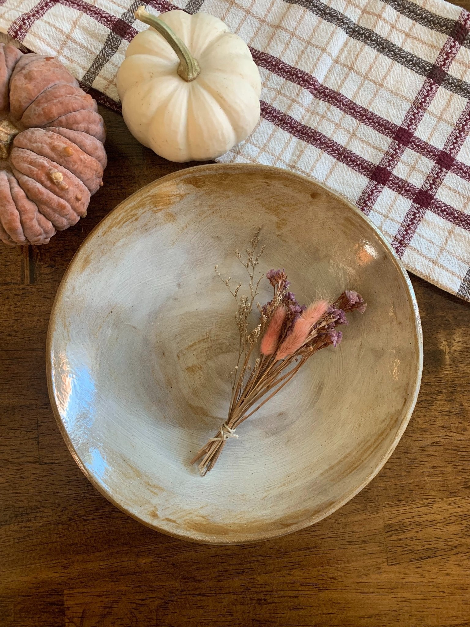 Decorative ceramic bowl with dried flowers on a wooden surface with pumpkins and a plaid cloth in the background.