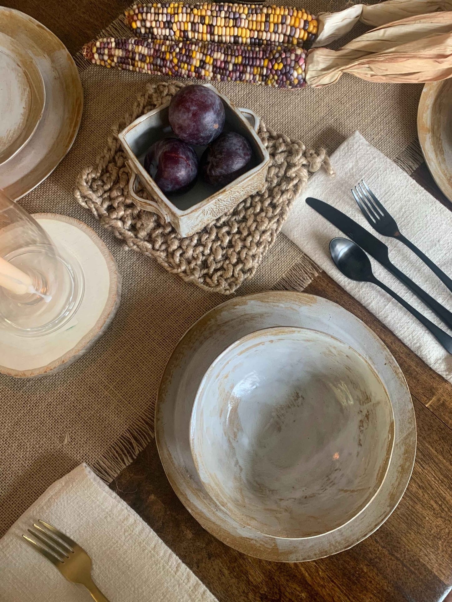 Dining table setting with handmade ceramic dinnerware, cutlery, and a small basket of plums on a textured tablecloth.