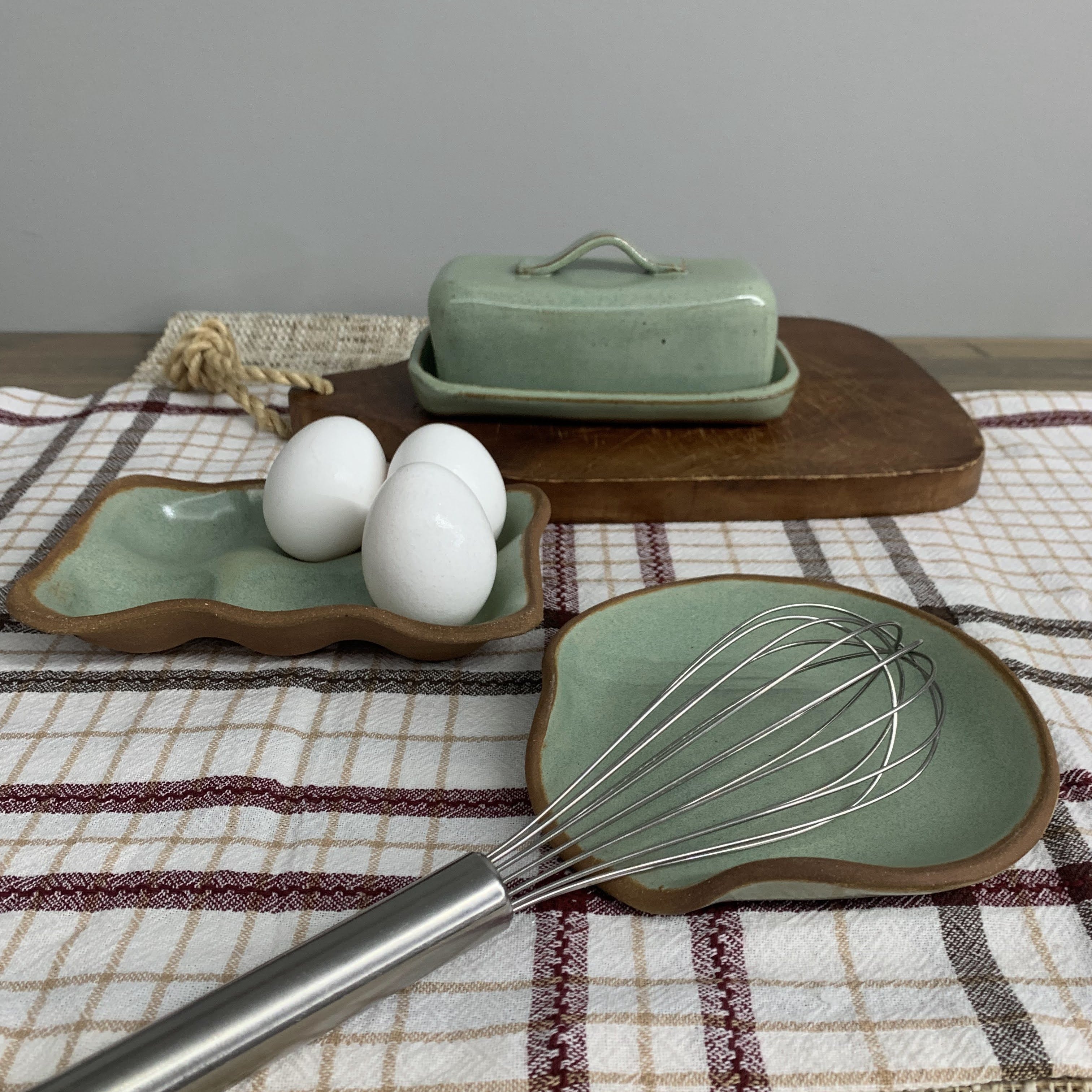 Green ceramic tray with eggs, butter dish, and whisk on a checkered tablecloth.