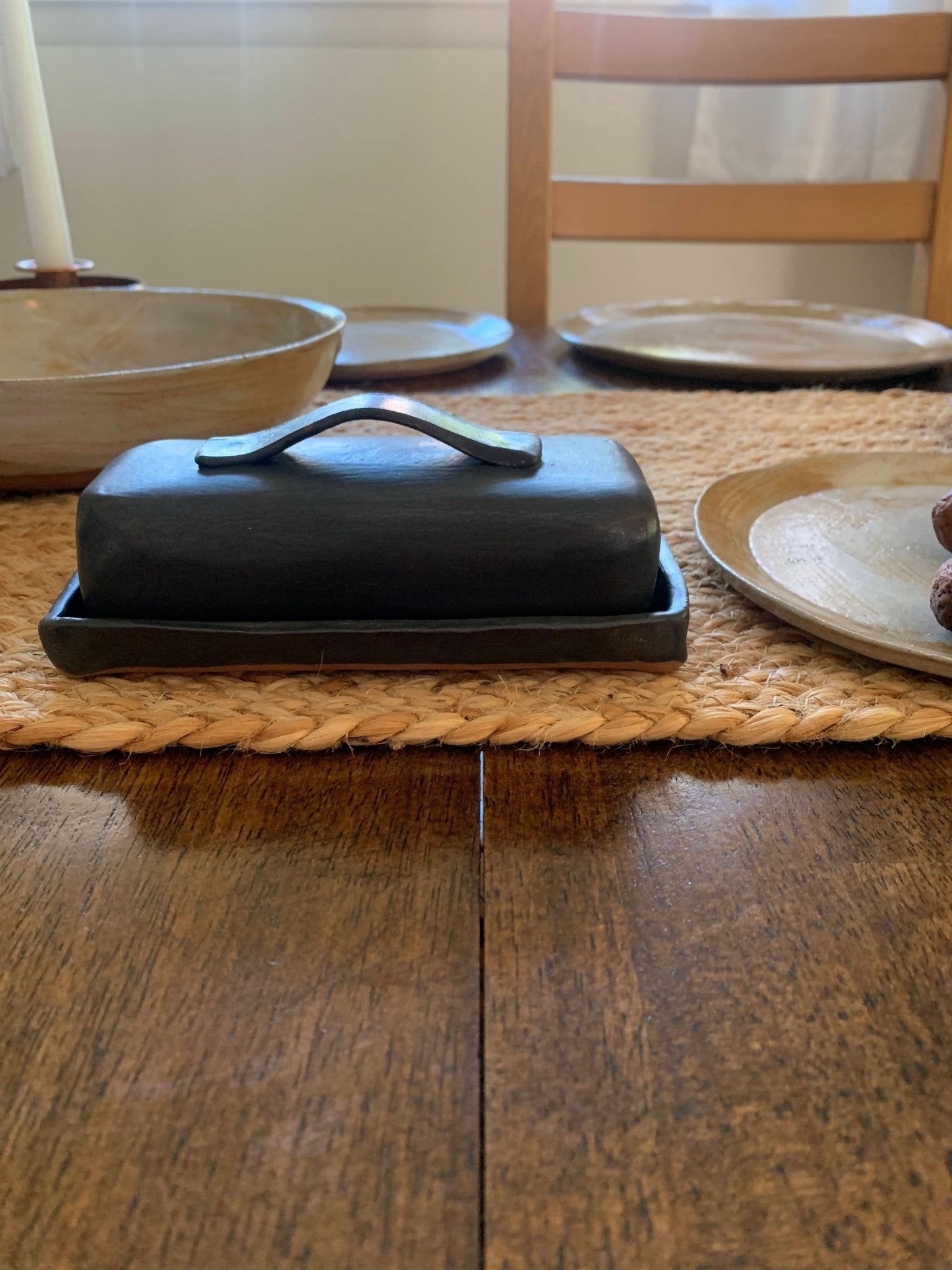 Black handcrafted butter dish on a wooden table with plates and bowls in the background