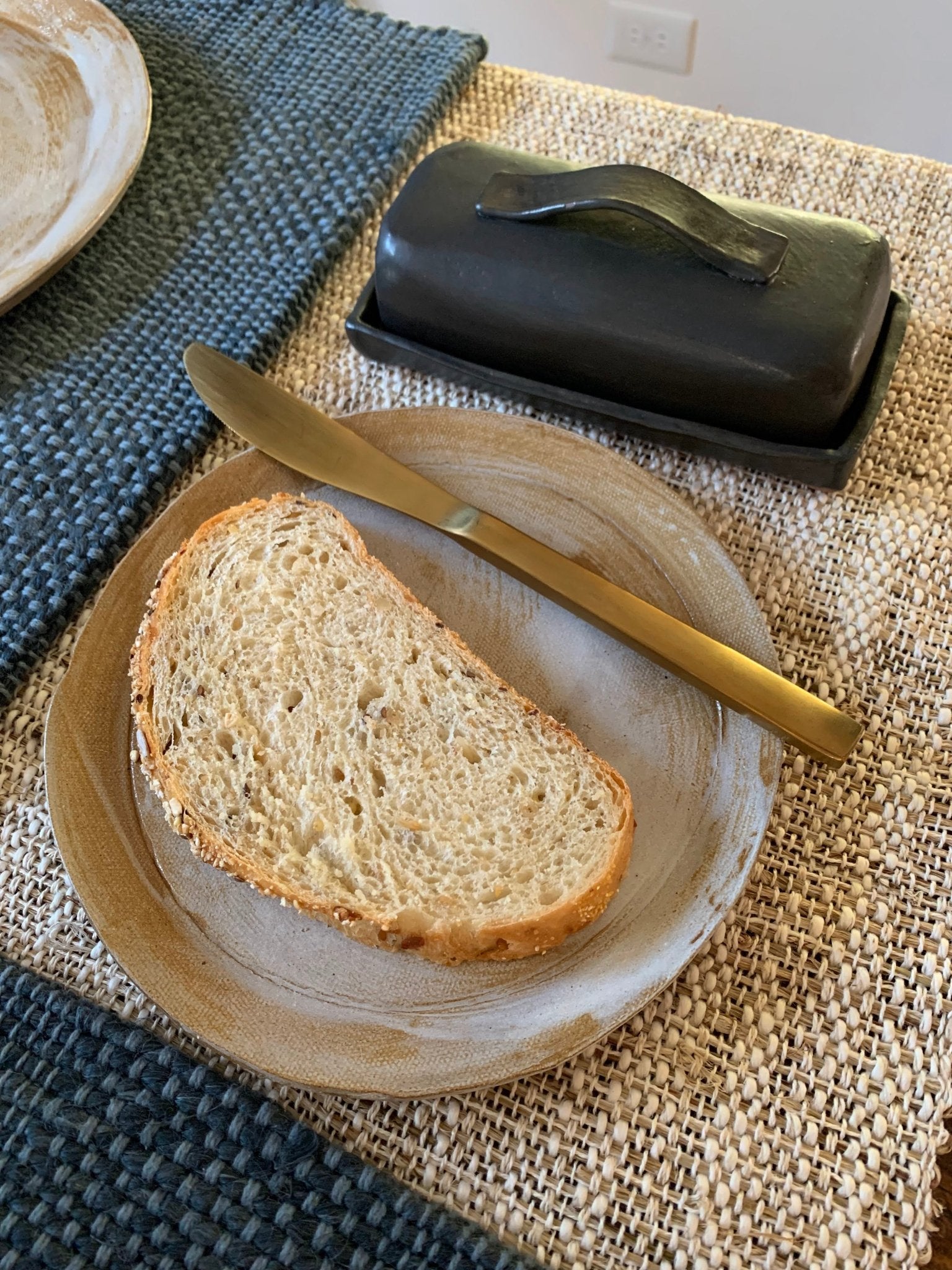 Slice of bread on a plate with a knife and stoneware butter dish on a textured surface