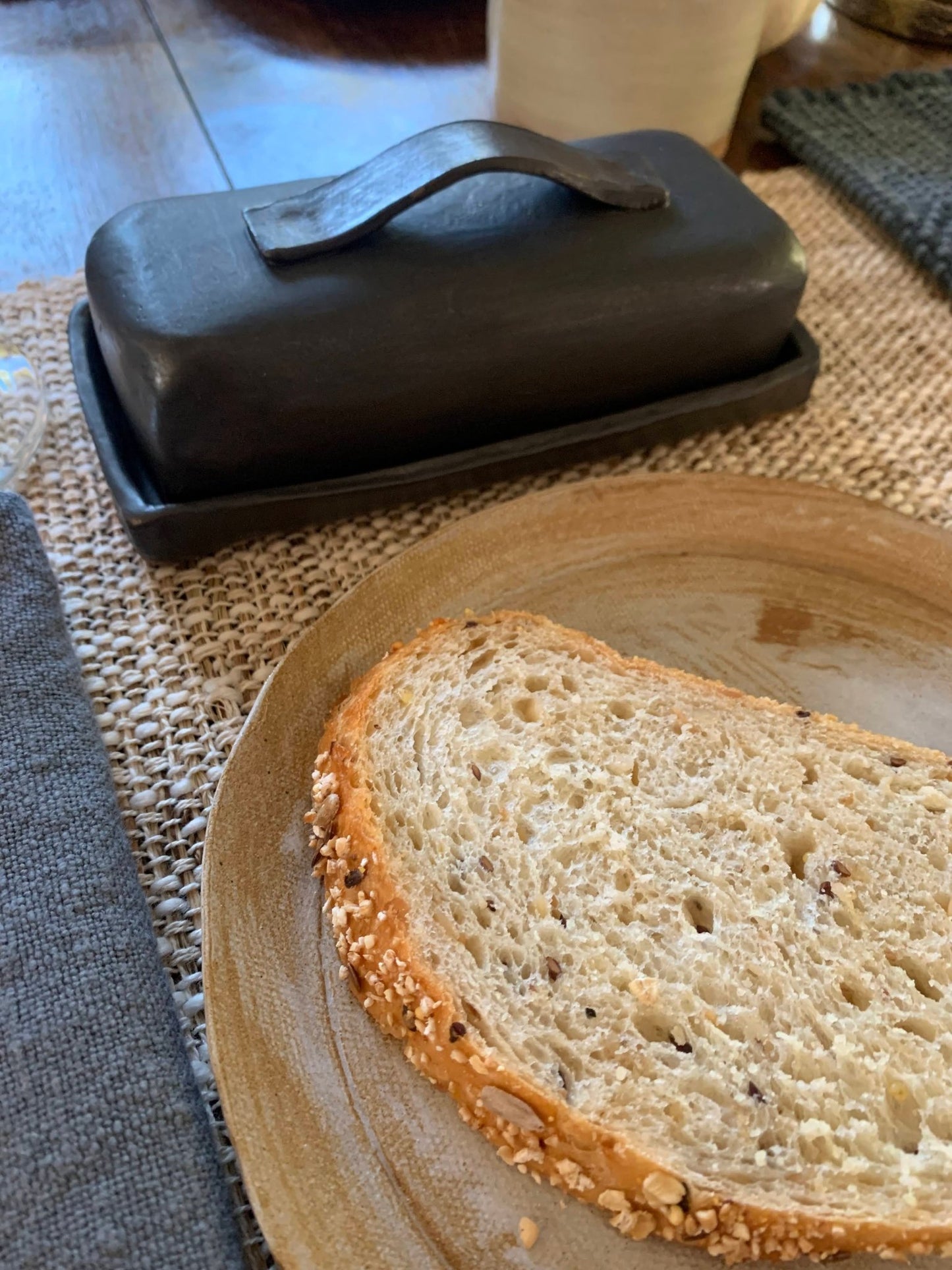 Slice of bread on a plate with a black ceramic handmade butter dish in the background