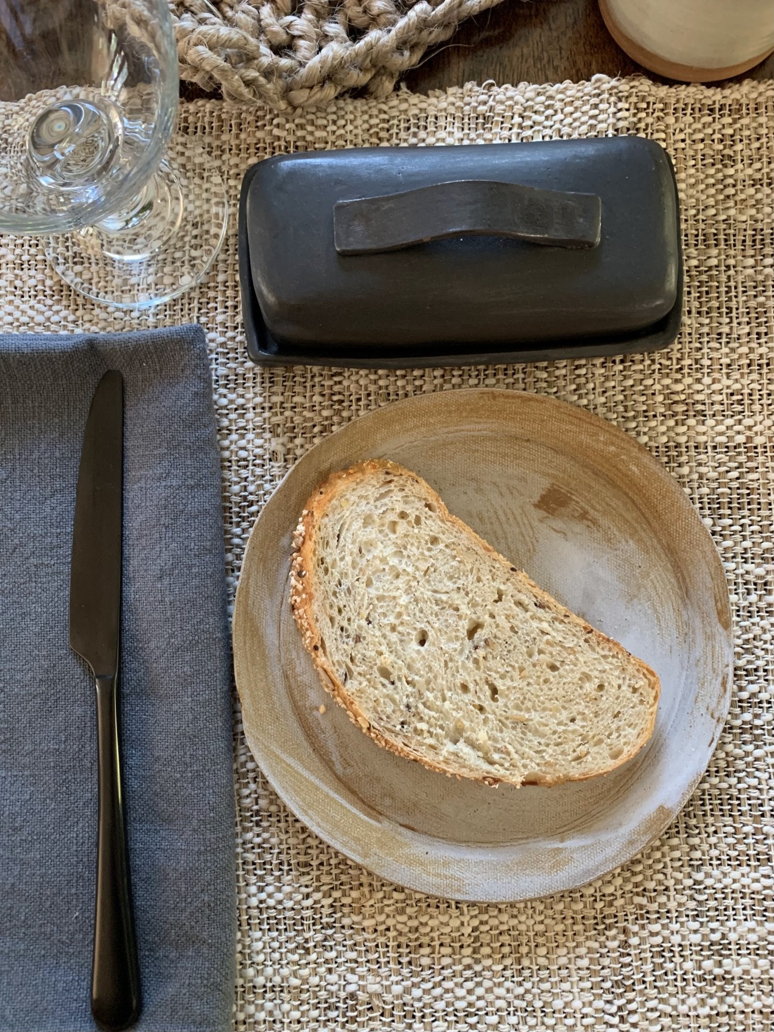 Sliced bread on a wooden plate with a knife and butter dish on a textured tablecloth.