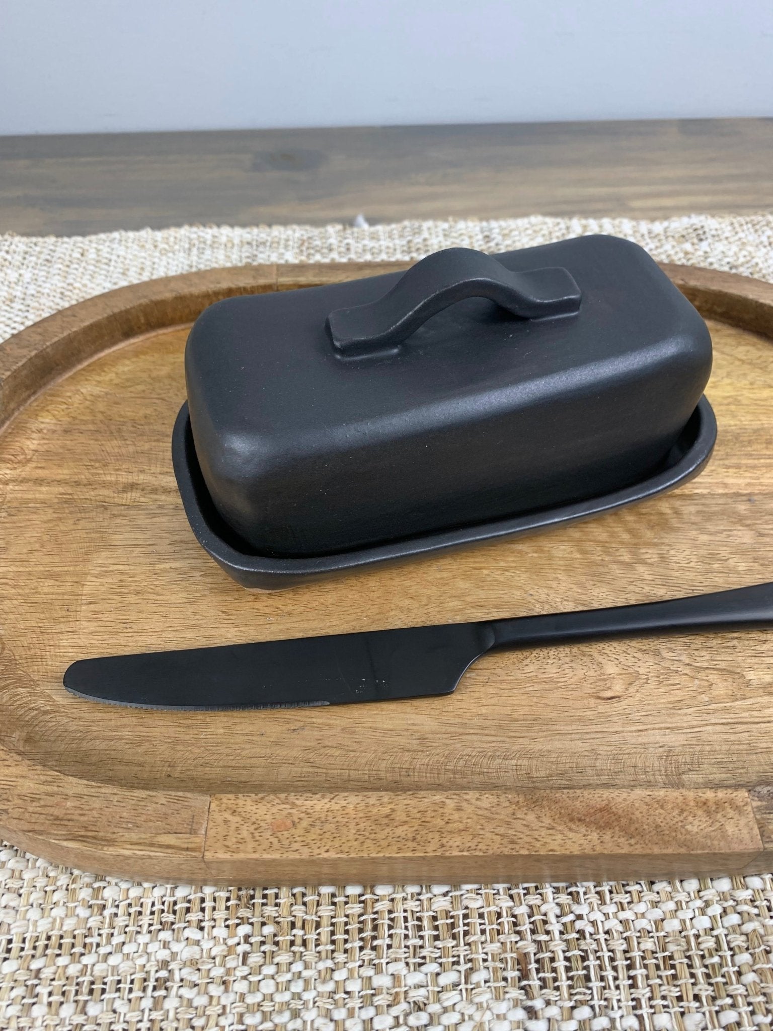 Black artisan made butter dish with lid and knife on a wooden tray with gray background