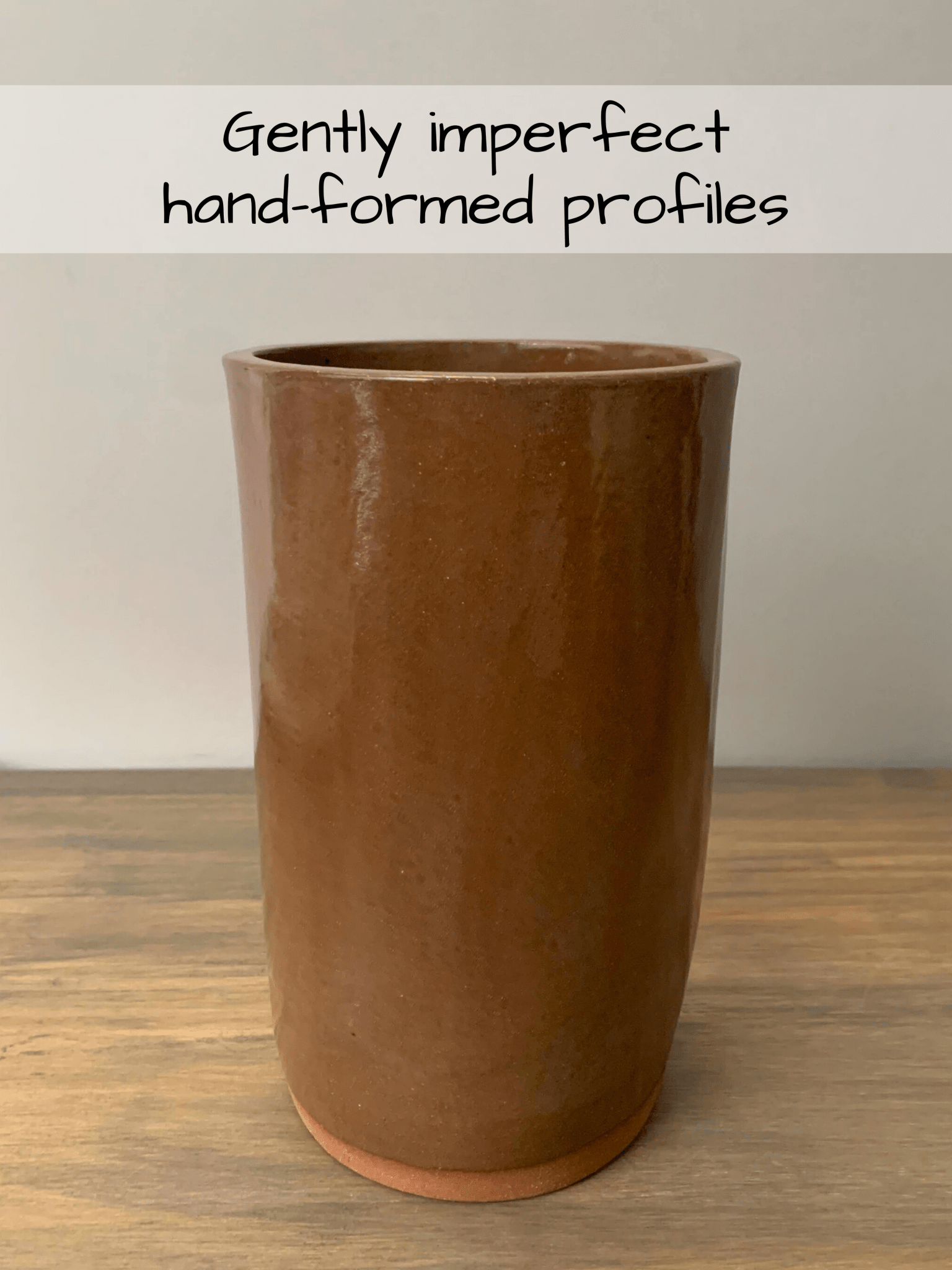Brown ceramic cylinder on a wooden surface with text 'Gently imperfect hand-formed profiles' above it.