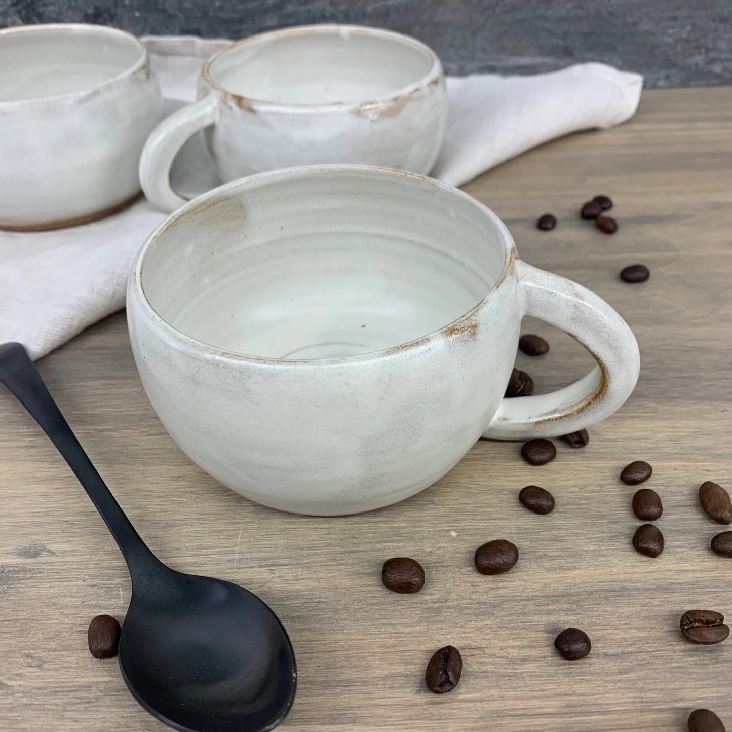 Two handmade ceramic coffee mugs on a wooden surface with scattered coffee beans and a black spoon.