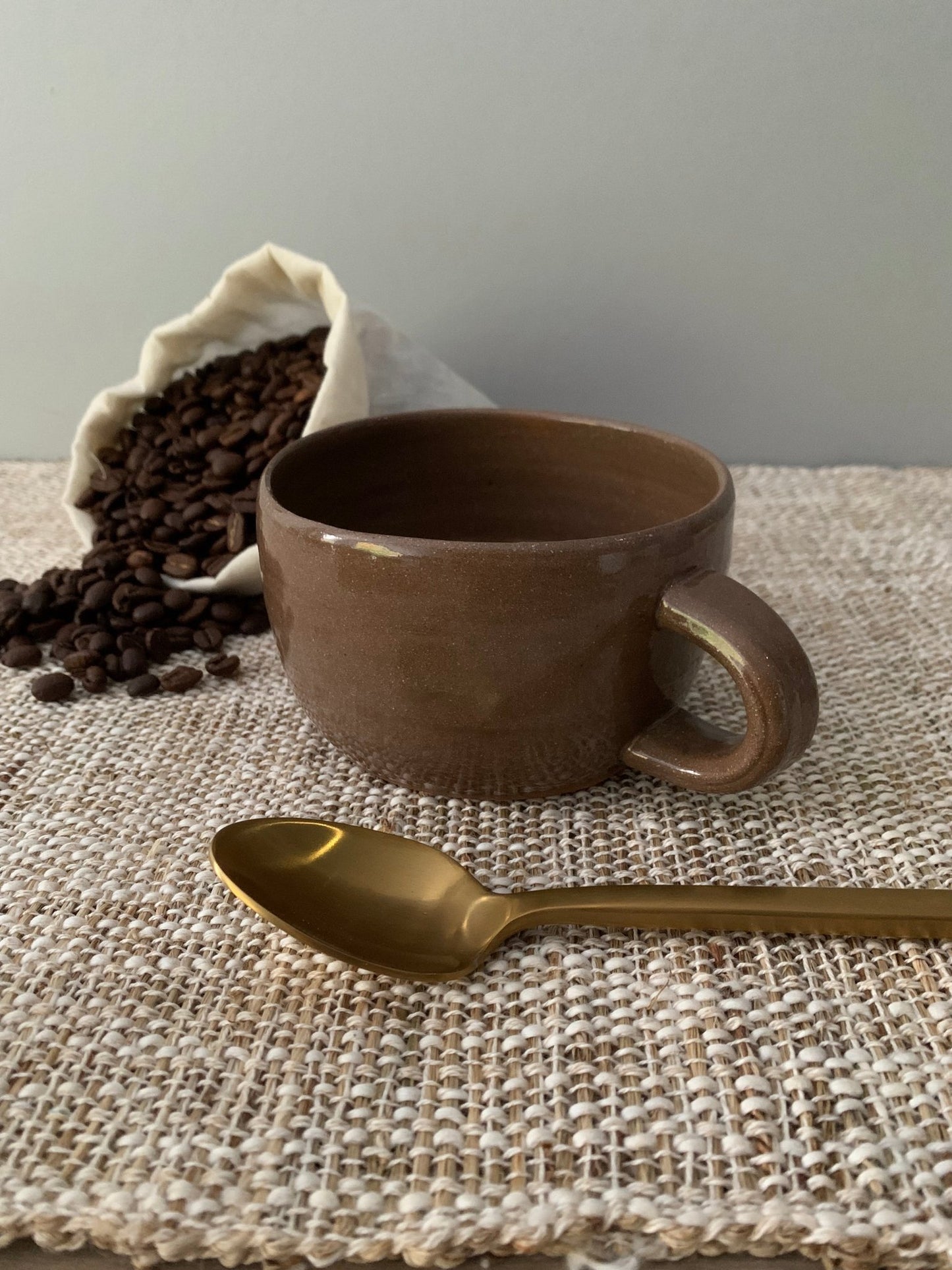 Brown ceramic mug with a gold spoon on a textured surface, with coffee beans and a white bag in the background.