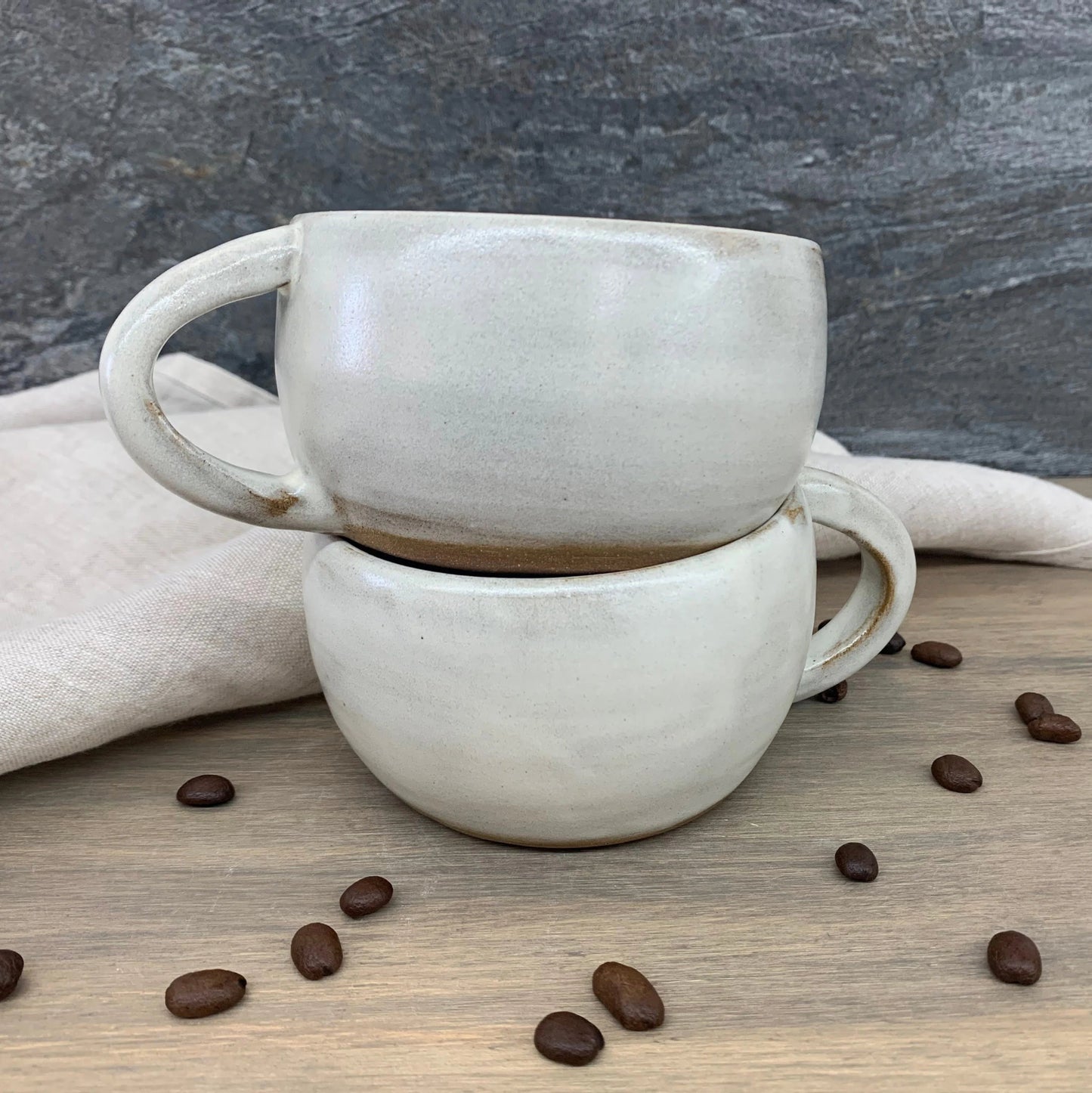 Two stacked handmade ceramic mugs on a wooden surface with scattered coffee beans.