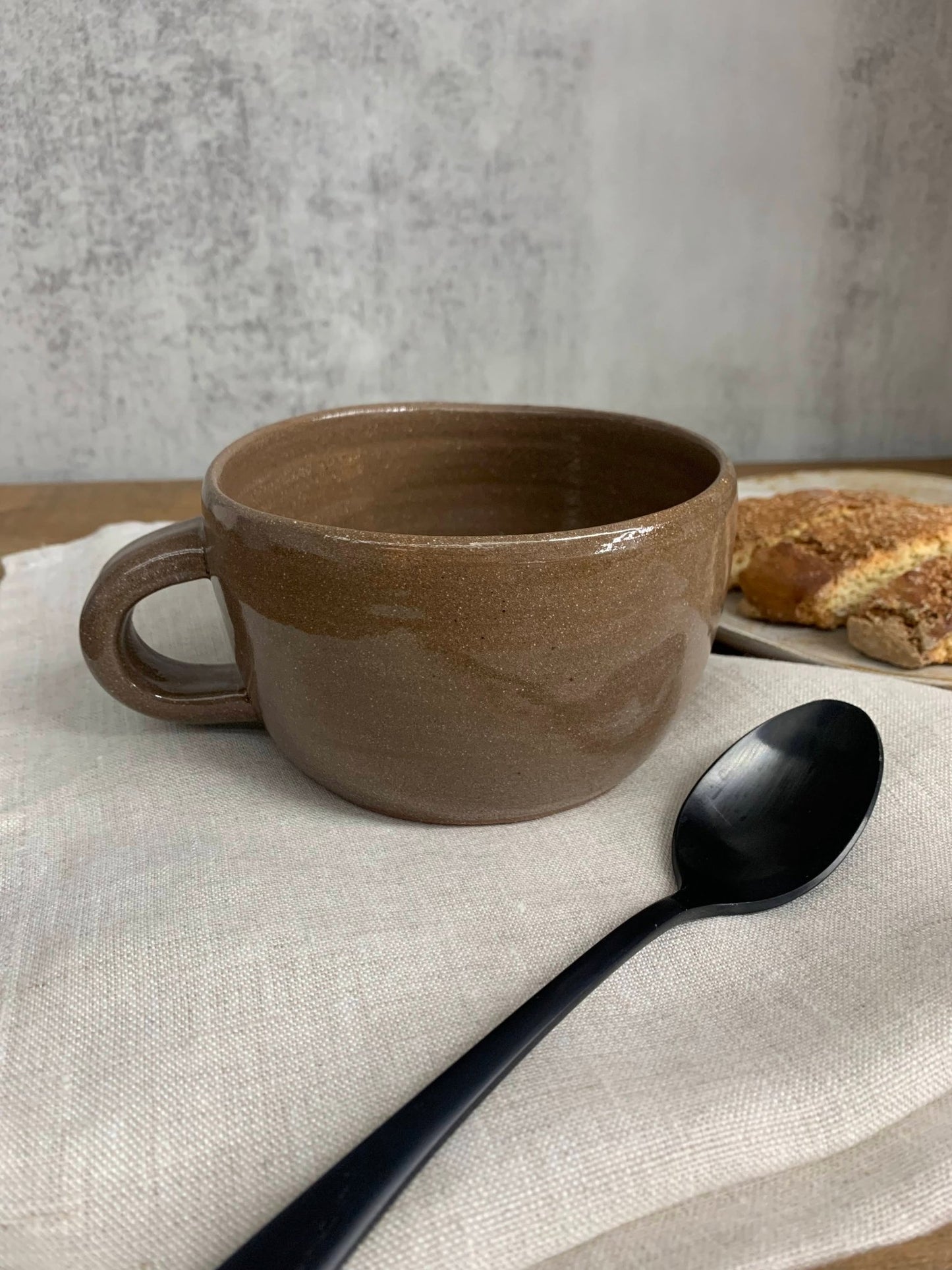 Brown ceramic mug with a black spoon on a light fabric surface, with a blurred background of bread and a plate.