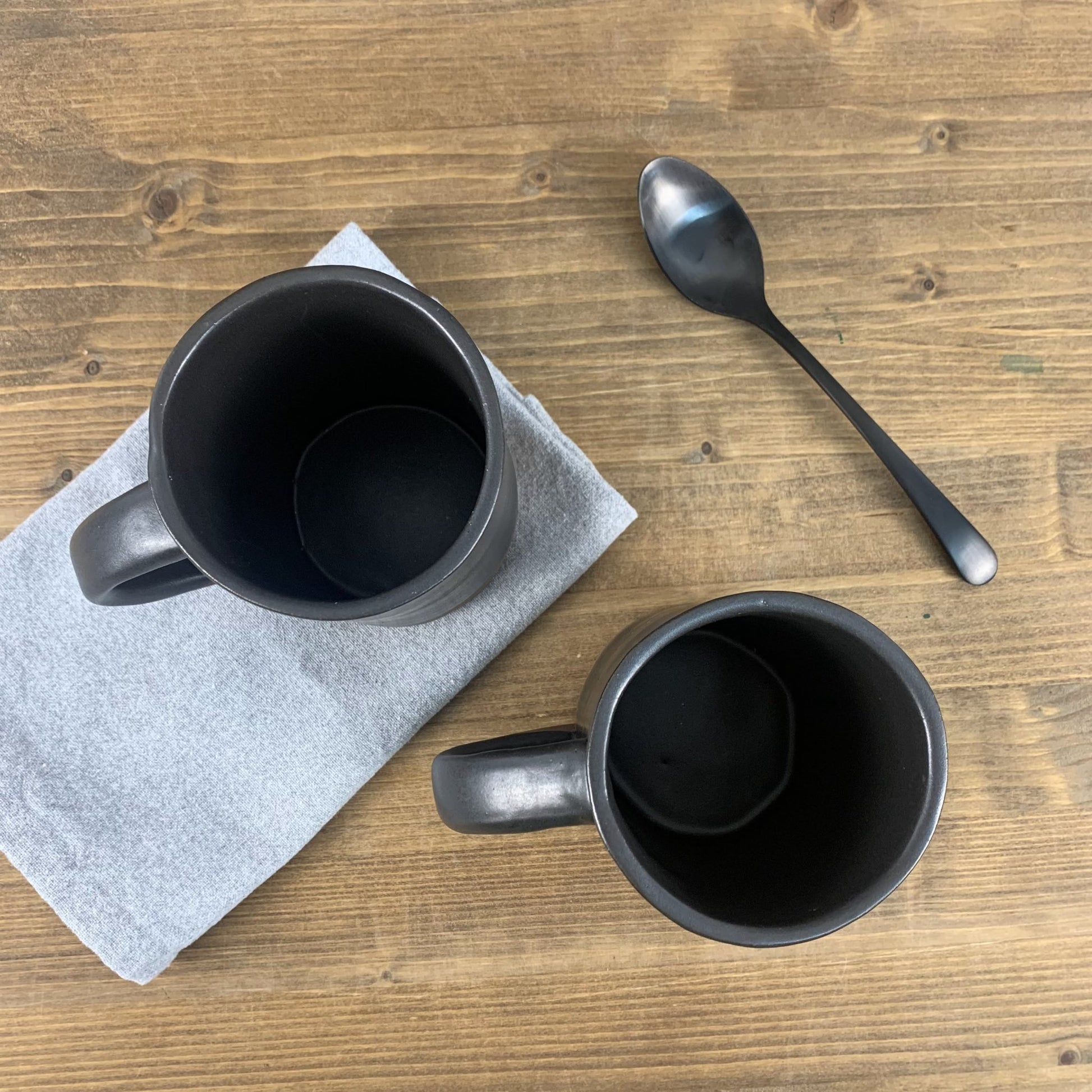 Two black stoneware mugs on a wooden table with a spoon and napkin.