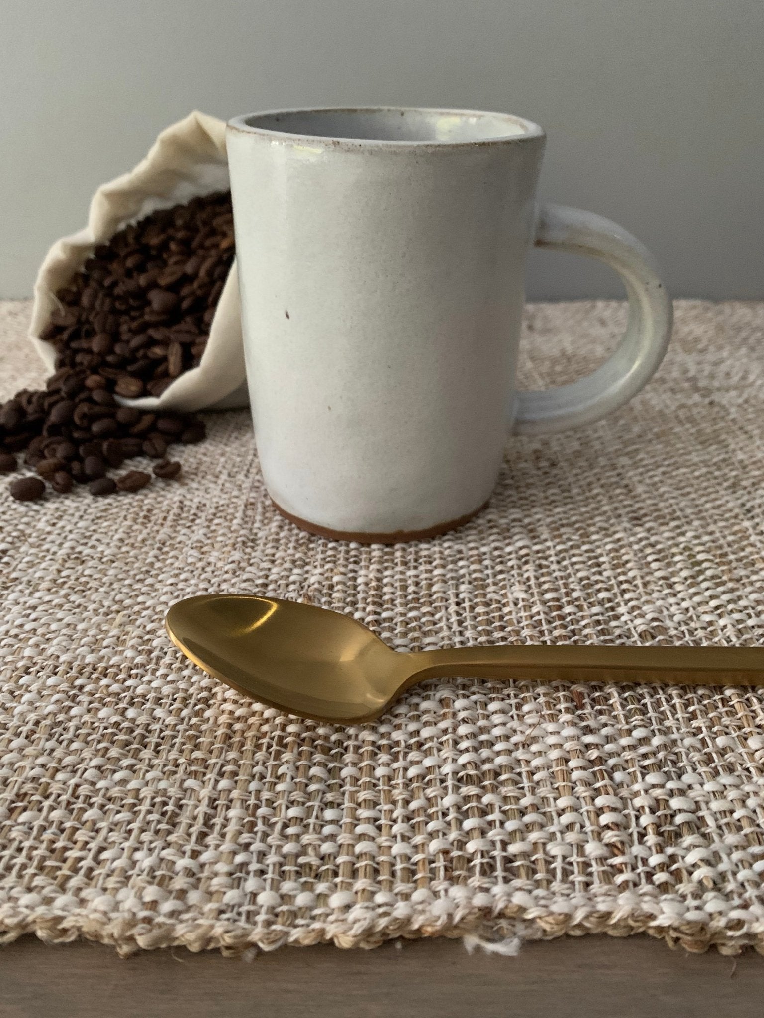 white ceramic mug with bag of coffee beans and a gold spoon on a textured place mat on table