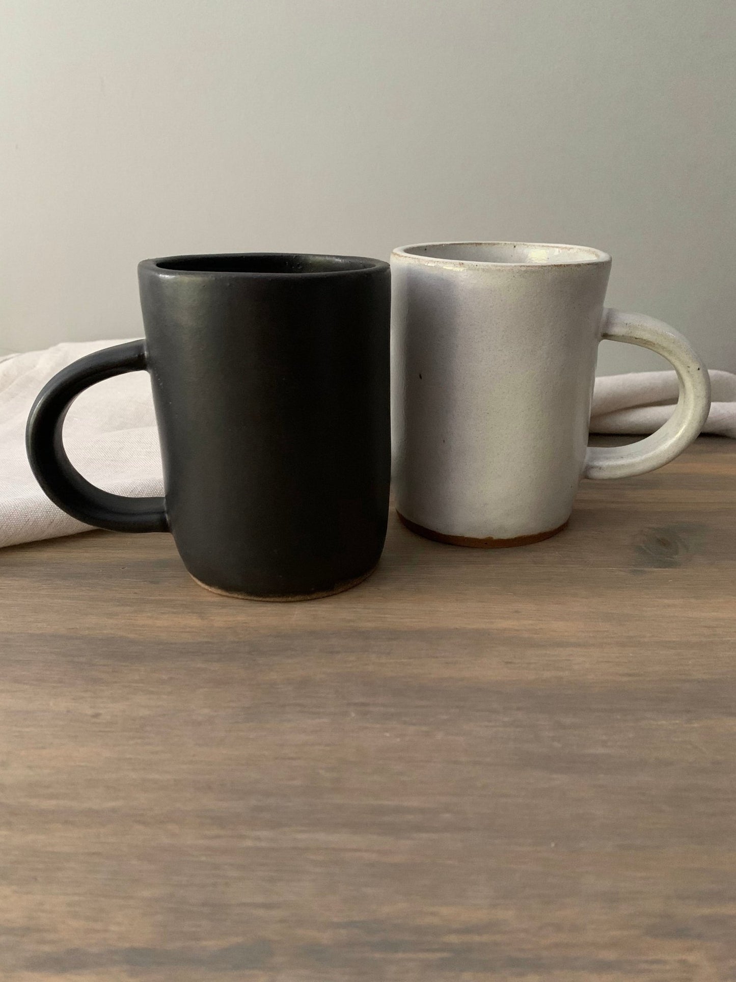Two handmade  ceramic mugs, one black and one gray, on a wooden surface with a neutral background.