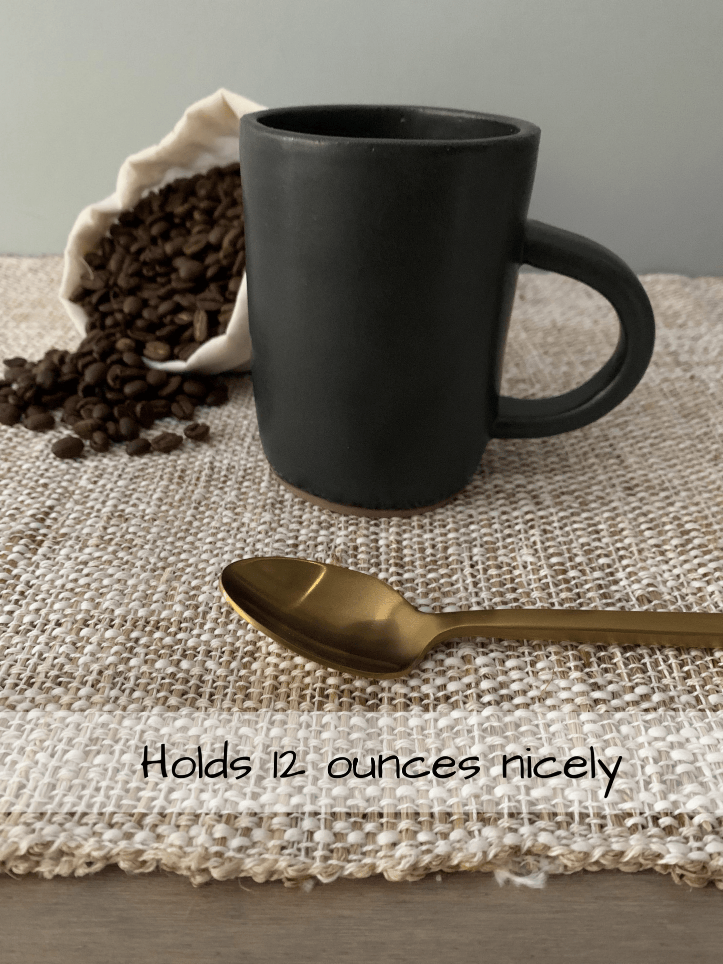 Black stoneware mug with a gold spoon and coffee beans on a textured surface