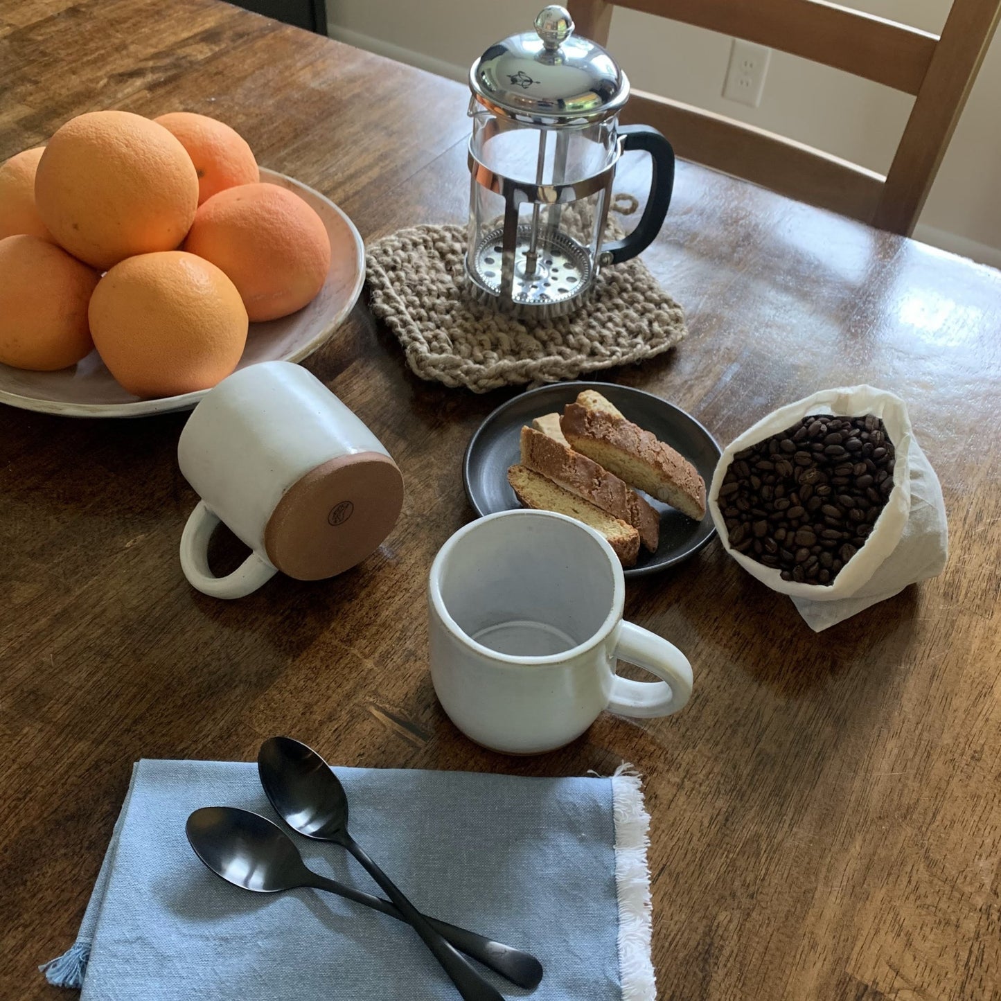 Table setting with oranges, coffee beans, handmade mugs, and a French press on a wooden table.