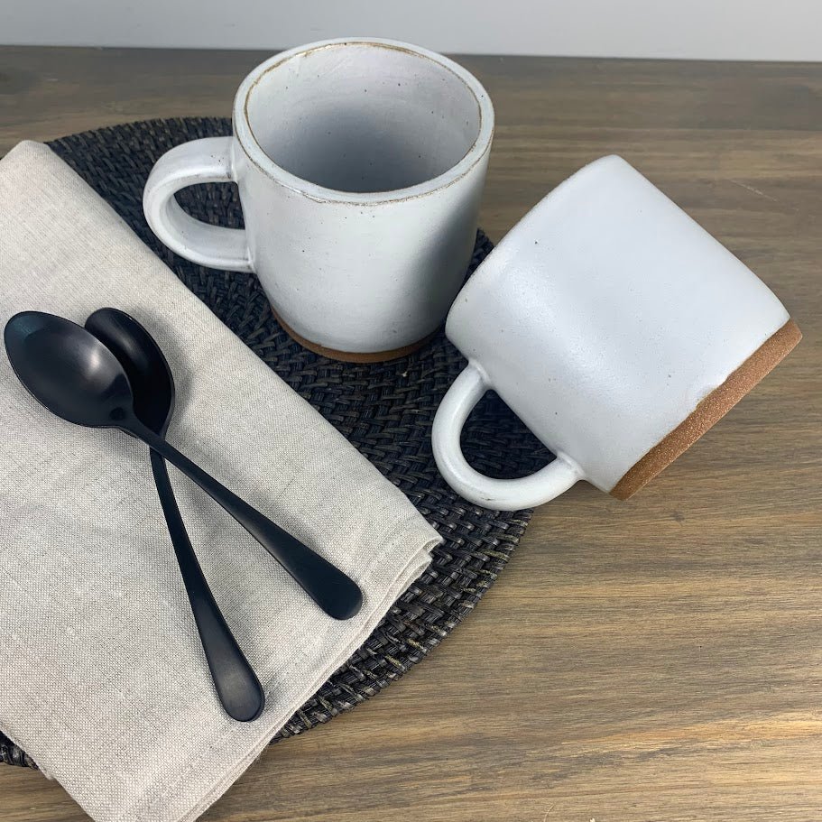 Two white handmade ceramic mugs with brown accents on a wooden table with black spoons and a beige napkin.