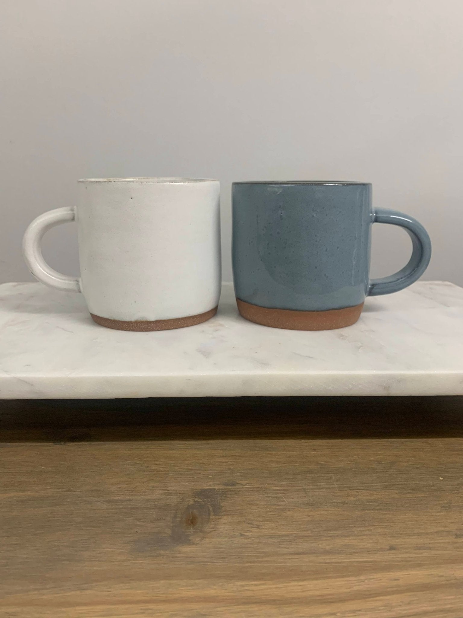 Two ceramic mugs, one white and one blue, on a marble surface with a wooden floor.
