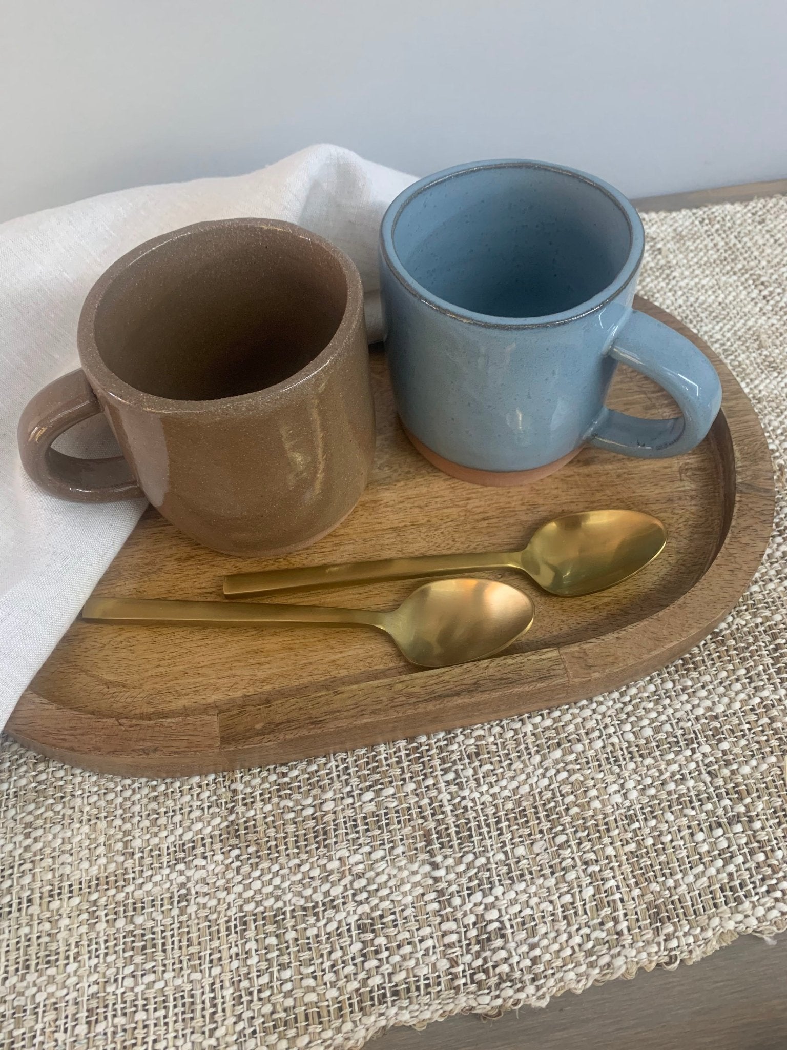 Two handmade  ceramic mugs on a wooden tray with gold spoons on a textured surface