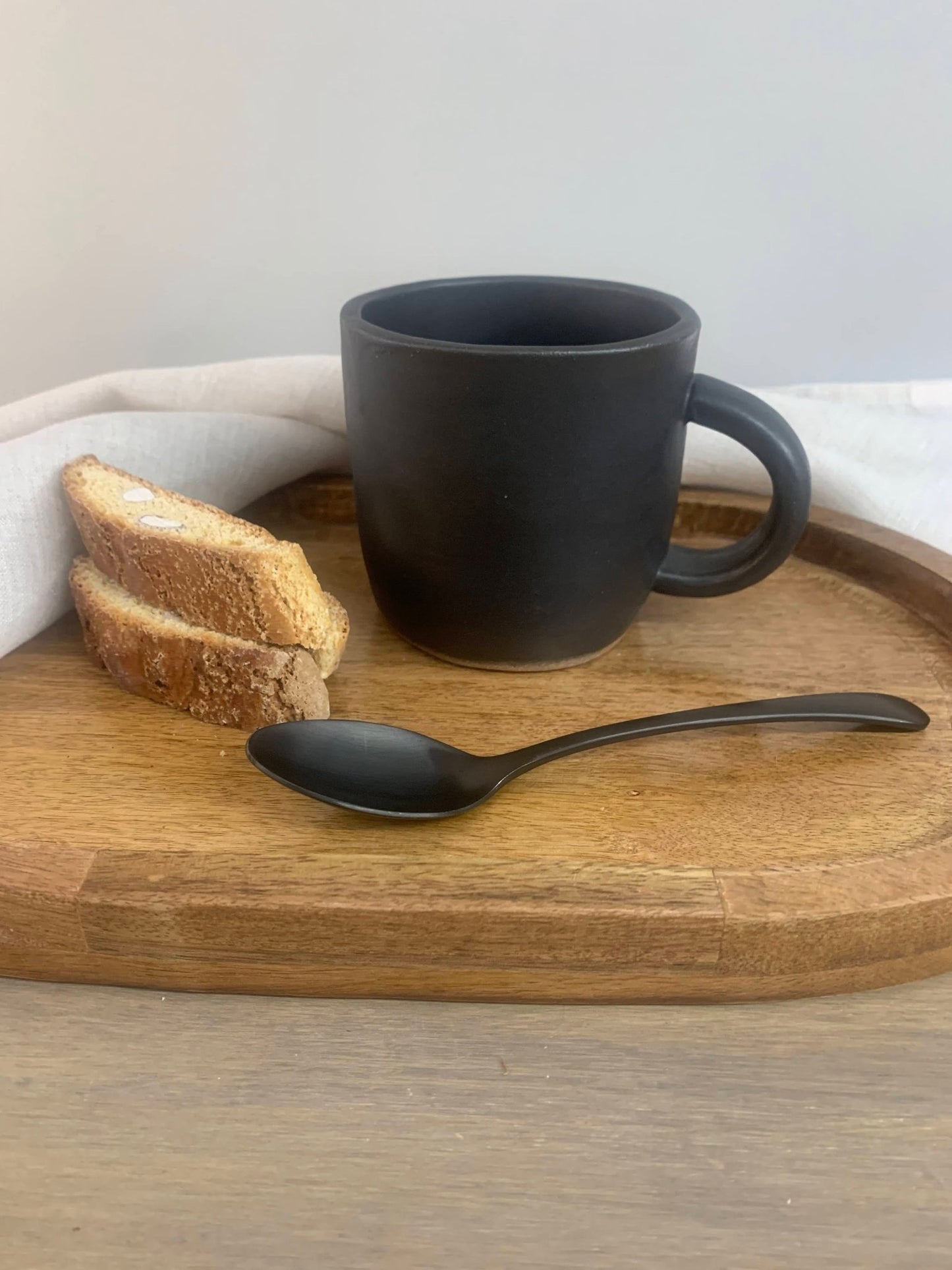 Black ceramic mug, spoon, and bread on a wooden tray with a white cloth
