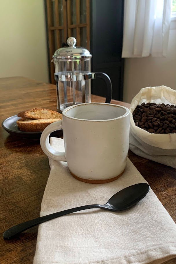 White handmade mug on a wooden table with a plate of toast, French press, and coffee beans.