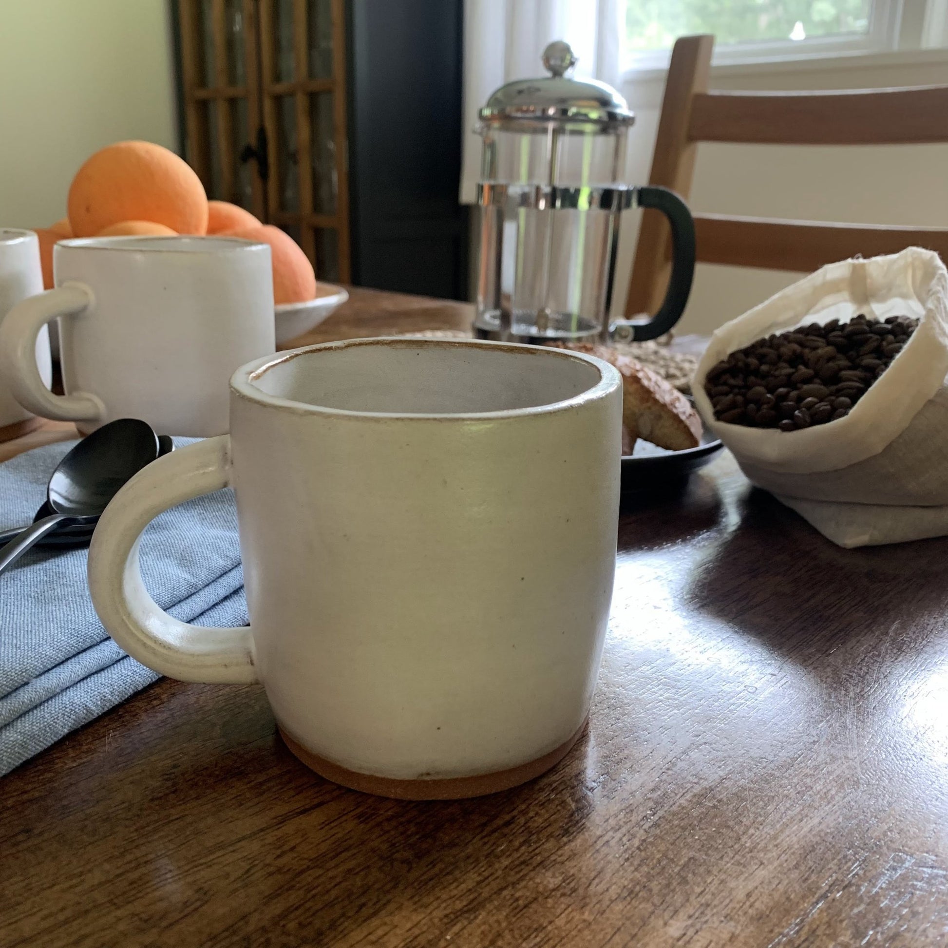White handmade ceramic mug on a wooden table with coffee beans, oranges, and a French press in the background.