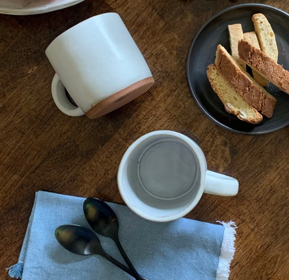 Two white handmade mugs on a wooden table with a plate of bread slices.