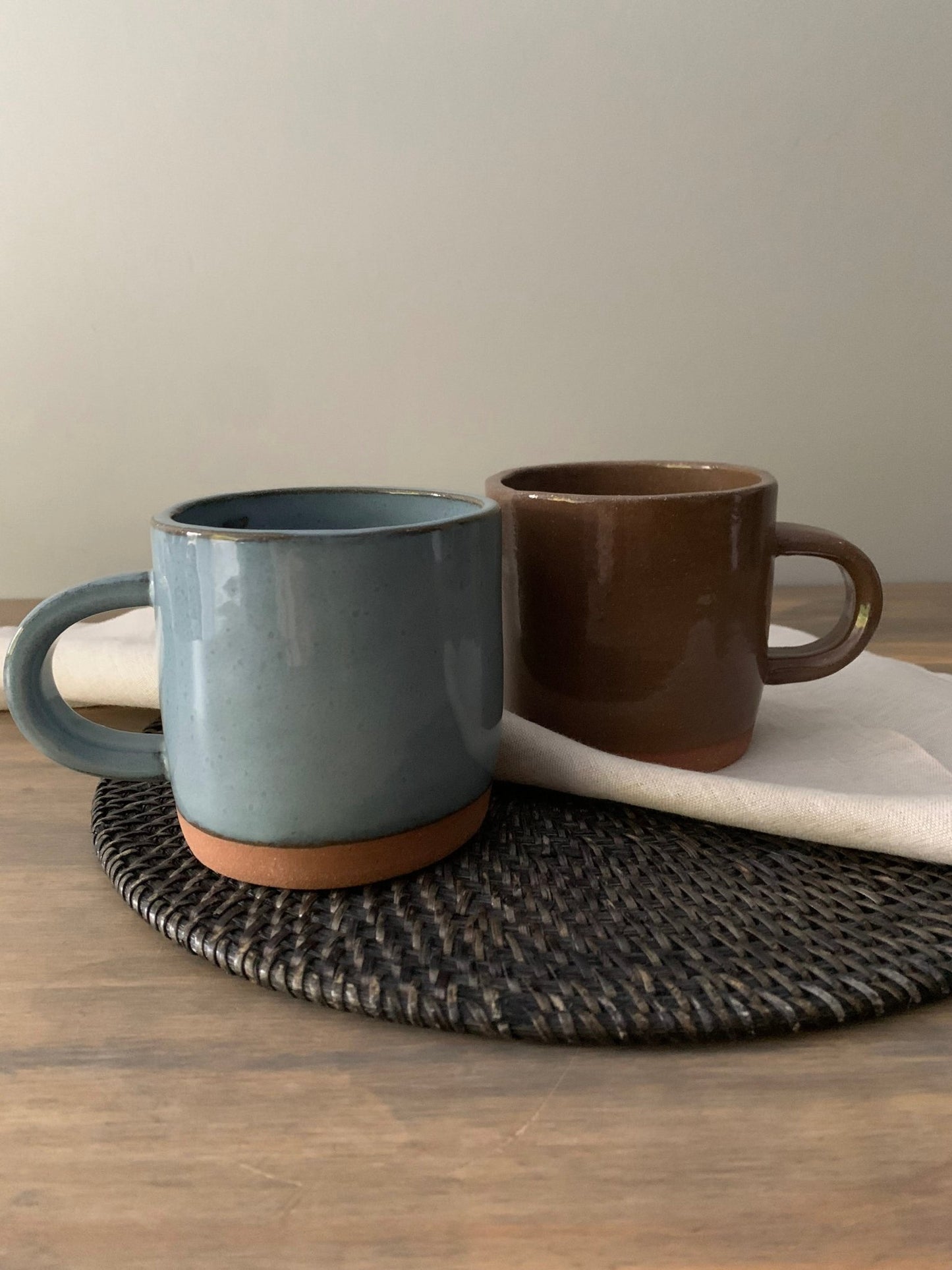 Two ceramic mugs, one blue and one brown, on a woven placemat with a neutral background.