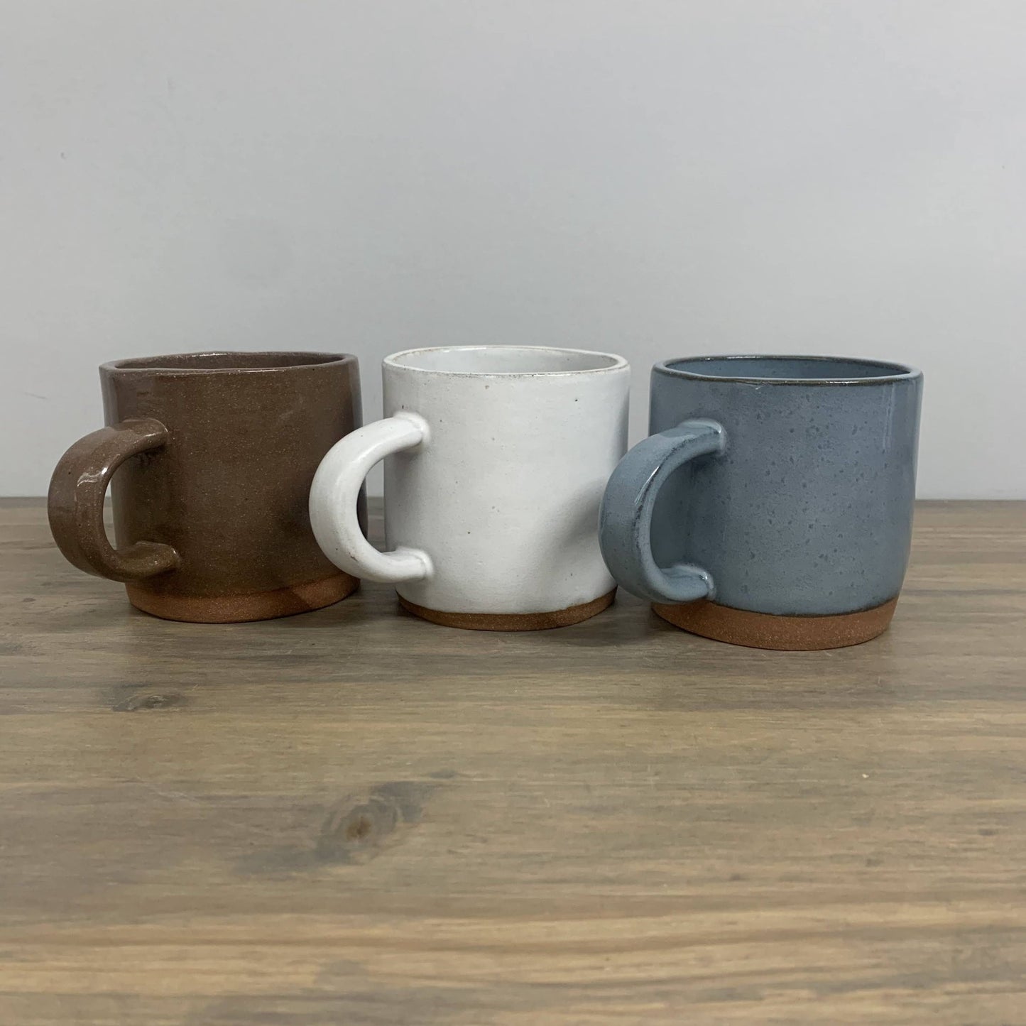 Three ceramic mugs in brown, white, and blue on a wooden surface with a gray background