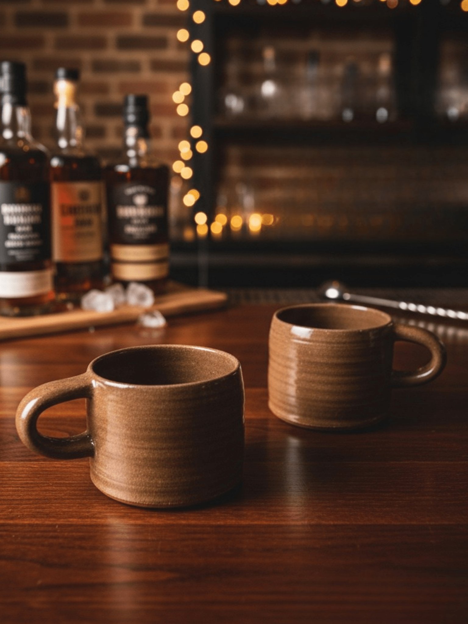 Two handmade  ceramic mugs on a bar counter with bottles and blurred lights in the background