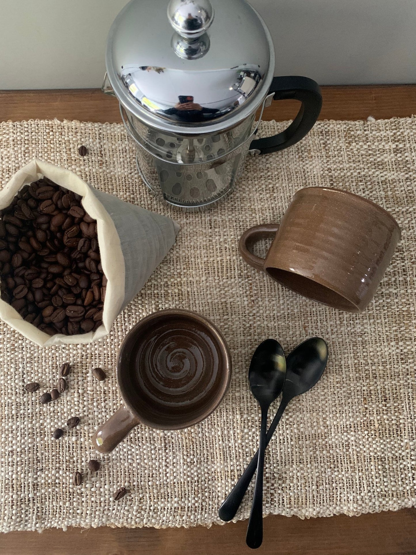 Coffee-making setup with French press, coffee beans, handmade mugs, and spoons on a textured surface.