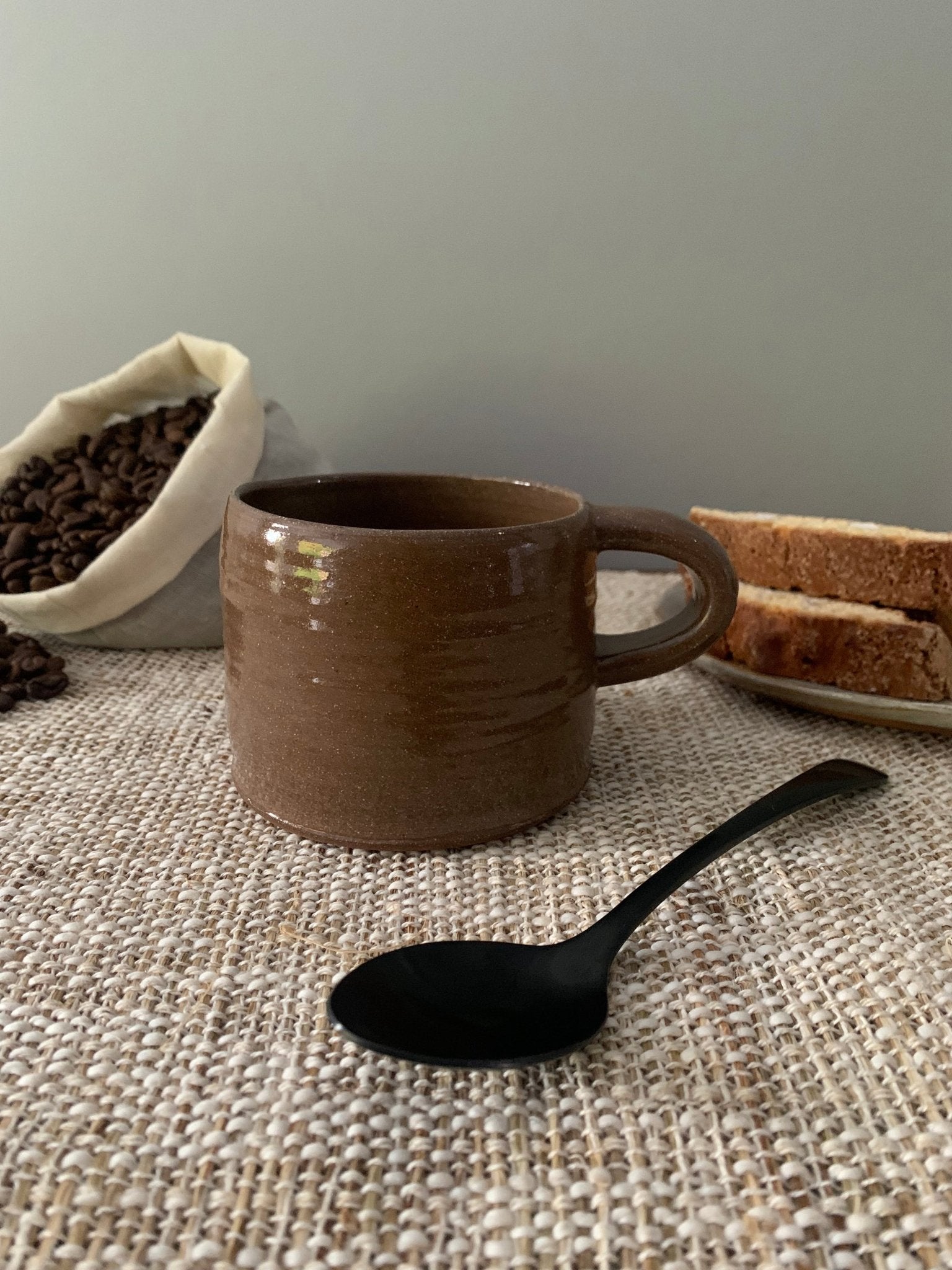 Brown ceramic mug with a black spoon on a textured surface, with coffee beans and bread in the background.