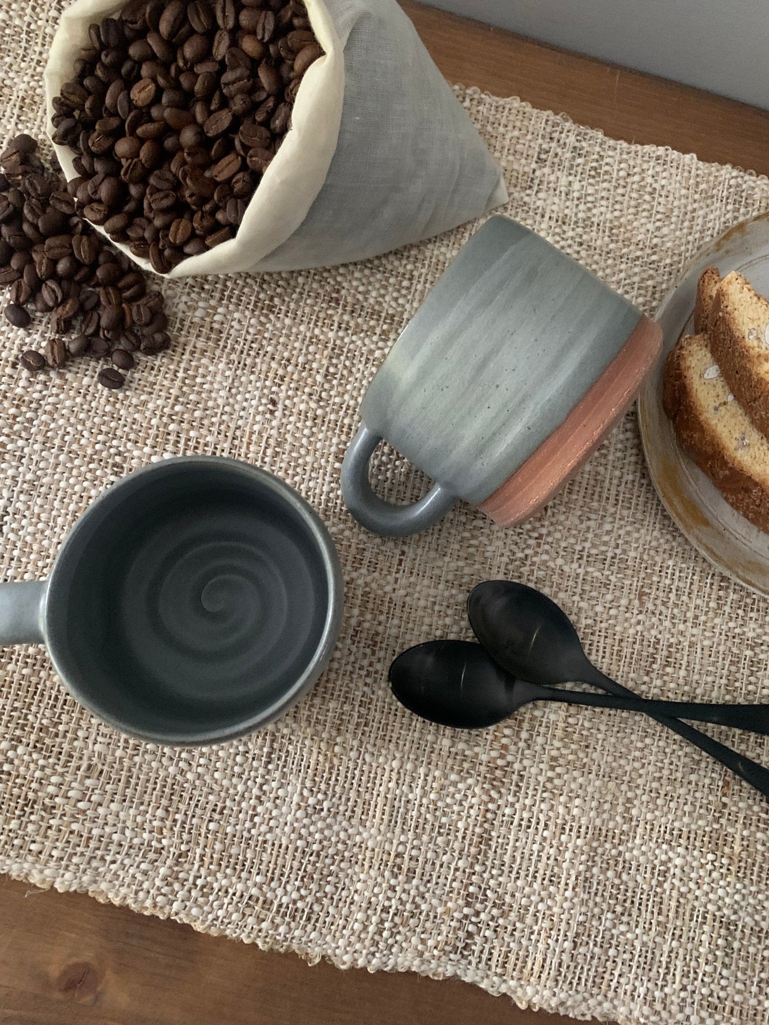 Two gray handmade ceramic mugs on a textured surface with coffee beans and a bowl of cookies.