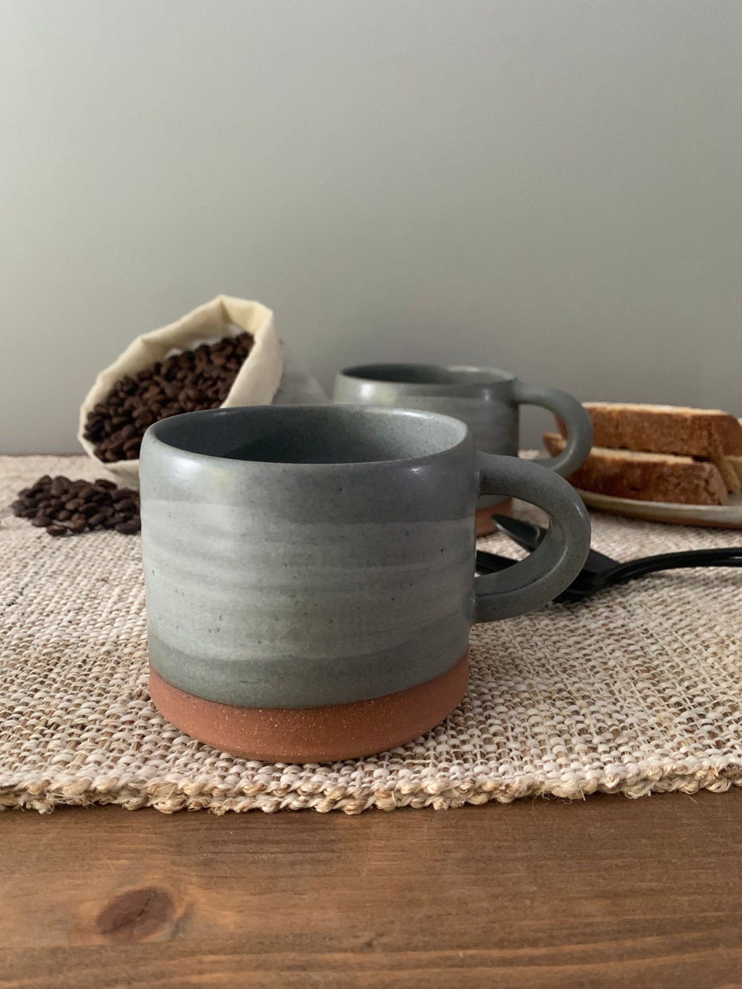 Two gray ceramic mugs on a textured surface with coffee beans and bread in the background.