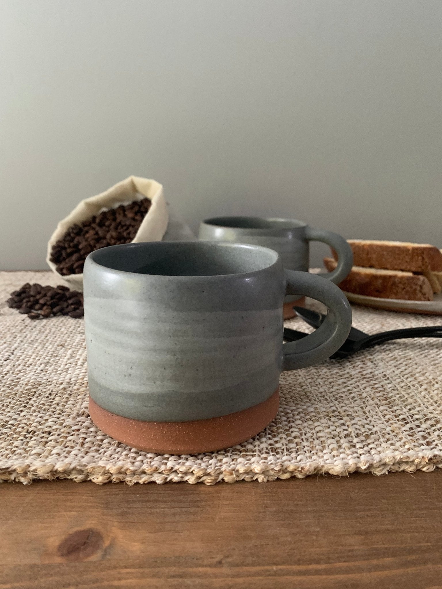 Two gray ceramic mugs on a textured surface with coffee beans and bread in the background.