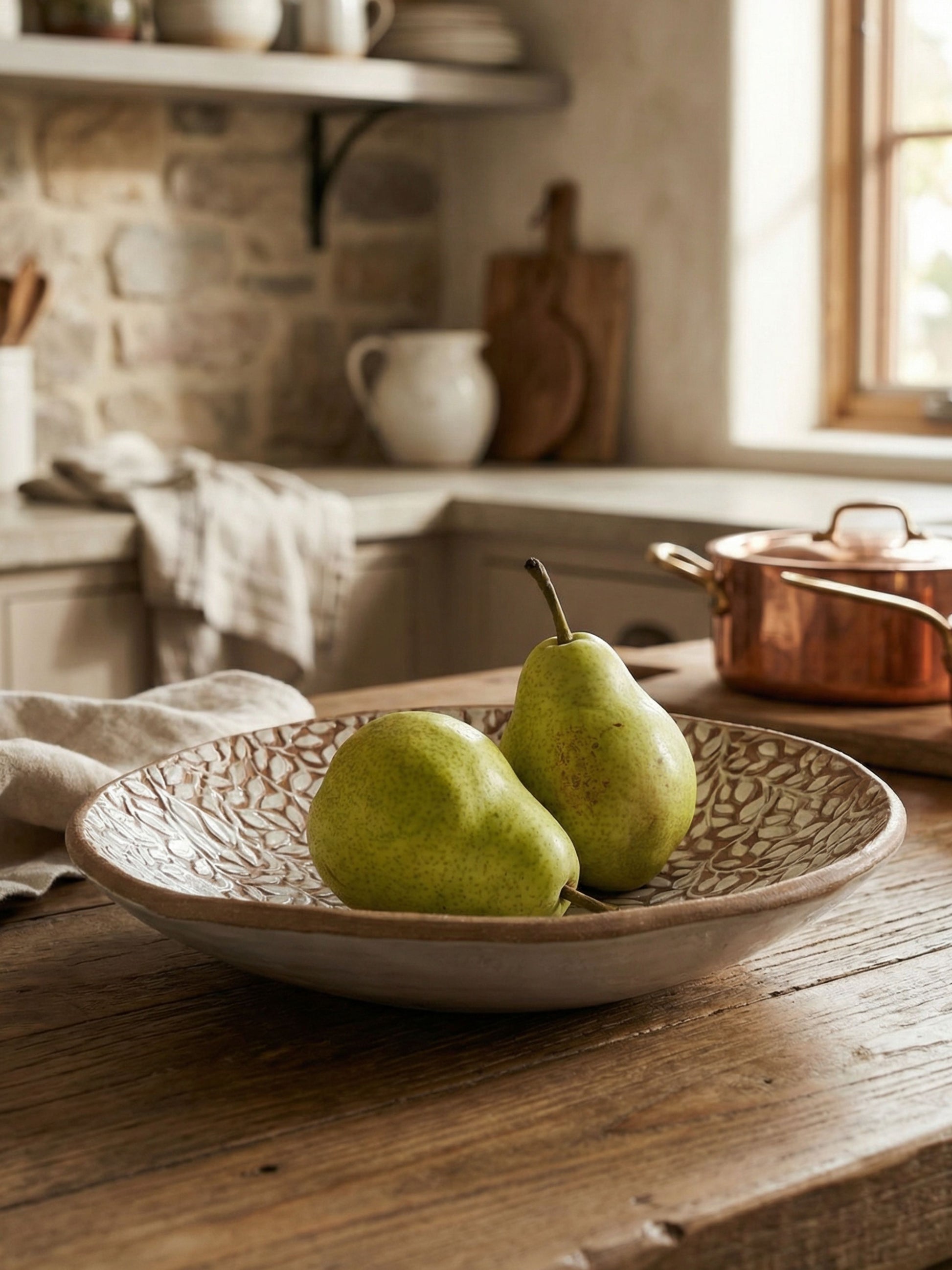 Two pears in a decorative handmade ceramic bowl on a wooden table with a kitchen background.
